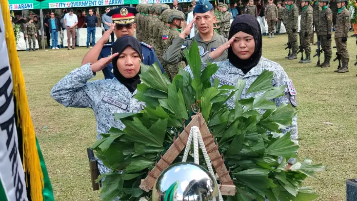 Uniformed military, air force, and police officers in headscarves render hand salutes behind a ceremonial green wreath at the Heroes Wall inside Kampo Ranao during the Marawi Liberation anniversary ceremony.