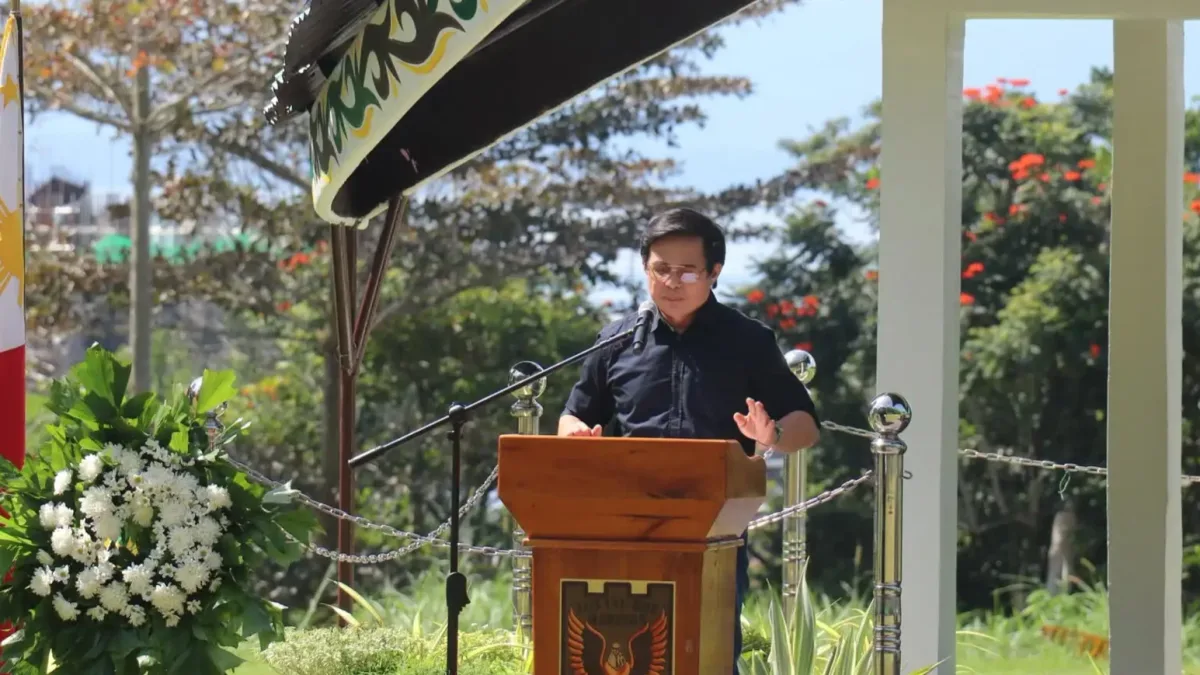 Marawi City Vice Mayor Majul U. Gandamra stands at a wooden podium under a covered outdoor pavilion flanked by floral arrangements while speaking during the ninth anniversary of Marawi's liberation.