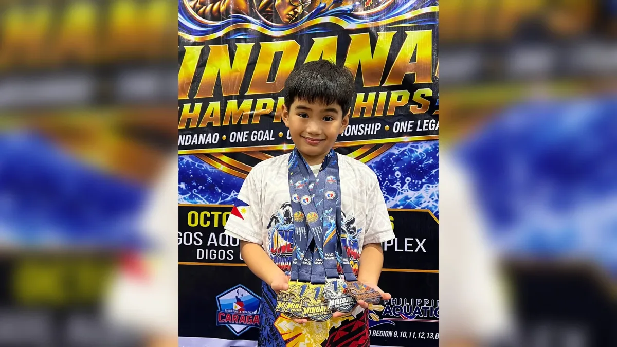 Seven-year-old swimmer Mike Constantine Pagaran displays three gold, one silver, and one bronze medal while standing in front of the official event backdrop at the One Mindanao Aquatic Championships 2026 in Digos City.