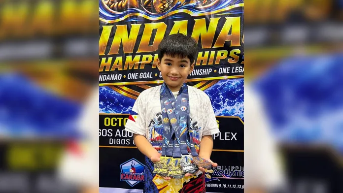 Seven-year-old swimmer Mike Constantine Pagaran displays three gold, one silver, and one bronze medal while standing in front of the official event backdrop at the One Mindanao Aquatic Championships 2026 in Digos City.