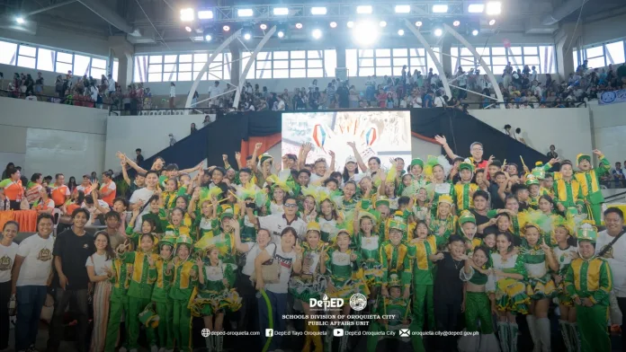 Members, majorettes, and teachers of Oroquieta City Central Elementary School's drum corps wearing bright green and yellow uniforms cheer and pose with their trophy inside an arena during the Inug-og Drum Corps Competition 2026.