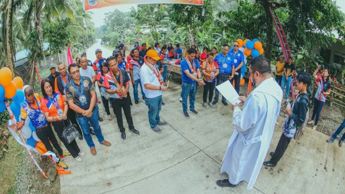 Local officials, DILG representatives, and residents gathering on a newly paved road while a priest conducts the blessing and turnover ceremony for a farm-to-market road and solar streetlights project in Barangay Agunod, Plaridel, Misamis Occidental.