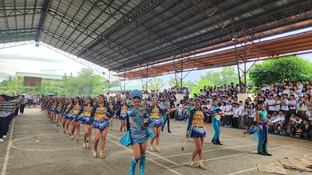 Majorettes and band dancers led by a head majorette in a blue and silver gown perform inside a covered court in front of cheering students during rehearsals for the 21st Dahunog sa Dipolog competition.