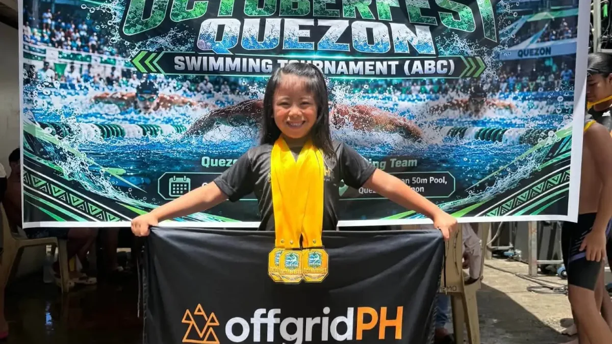 Six-year-old swimmer Solana Aislah "Daday" Ayco smiles as she holds up a sponsor flag while wearing five gold medals in front of the Oktoberfest Quezon Swimming Tournament banner.