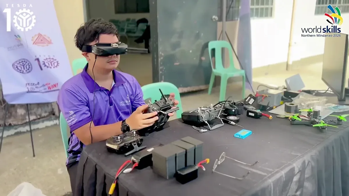 Technical-vocational trainee Miguel S. Estenzo wears FPV goggles and holds a radio controller as he demonstrates drone flight simulation technology at a WorldSkills Philippines display desk.
