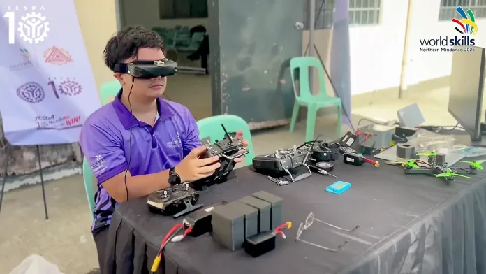 Technical-vocational trainee Miguel S. Estenzo wears FPV goggles and holds a radio controller as he demonstrates drone flight simulation technology at a WorldSkills Philippines display desk.