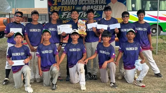 Student-athletes and coaches of the Valencia National High School baseball team, wearing blue jerseys with "Valencia City" branding, display their secondary division championship trophy, certificates, and cash prize envelopes following the 1st Davao Occidental Softball and Baseball Festival.