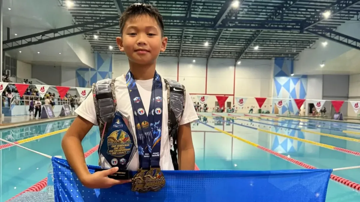 Nine-year-old swimmer Xav Roblin Galeon of Cagayan de Oro holds his Most Outstanding Swimmer trophy, wears five gold medals, and displays a sponsor banner poolside at the One Mindanao Aquatic Championships 2026 in Digos City.