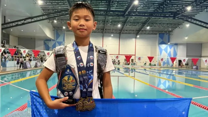 Nine-year-old swimmer Xav Roblin Galeon of Cagayan de Oro holds his Most Outstanding Swimmer trophy, wears five gold medals, and displays a sponsor banner poolside at the One Mindanao Aquatic Championships 2026 in Digos City.