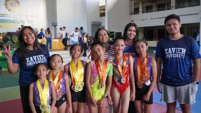 Young female gymnasts wearing competition leotards and medals pose with their coaches, clad in Xavier University blue shirts, following their medal haul at the Cagayan de Oro Division Meet.