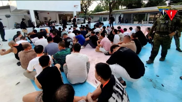 Dozens of foreign nationals seated on the floor of an outdoor court area while Philippine military personnel from Joint Task Force ZAMPELAN guard the perimeter during an anti-scam operation in Zamboanga Sibugay.